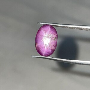 May include: A polished, oval-shaped, purplish-pink gemstone is held by a pair of silver tweezers. The gem has a star-like effect in the center. The background is a soft gray.