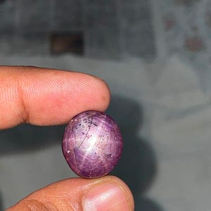 May include: A round, cabochon-cut star ruby gemstone with a deep purplish-red hue. The stone features a distinct six-rayed star effect, known as asterism, visible across its polished surface, along with natural dark inclusions within the crystal.