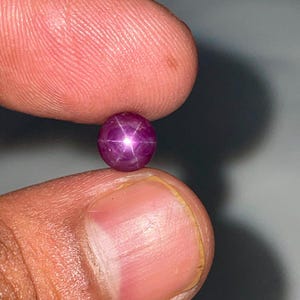 May include: A round, cabochon-cut star ruby gemstone displaying a distinct six-rayed star effect, known as asterism, centred on its deep magenta-red surface. The gemstone is polished to a smooth, glossy finish, reflecting light from its curved top.