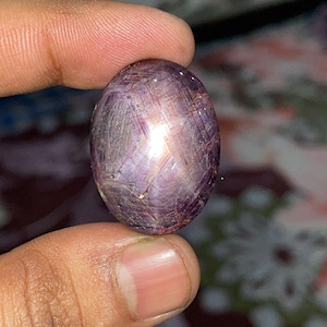 May include: Oval-shaped, purplish-red gemstone with a star-like effect. The gem is held between fingers, showcasing its polished surface and internal light play. The stone's color ranges from deep red to violet, with visible inclusions.