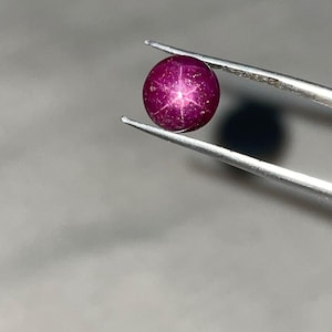 Puede incluir: Una piedra preciosa de rubí estrella de color magenta intenso y forma redonda, que presenta un patrón de estrella de seis puntas en su superficie. La piedra tallada en cabujón está sujeta por unas pinzas metálicas de tono plateado.