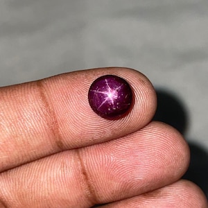 Pode incluir: Uma pedra preciosa de rubi estrela de corte cabochão redondo com uma cor vermelho magenta profundo. A pedra apresenta um efeito de estrela de seis raios distinto e centrado, conhecido como asterismo, que aparece como um reflexo branco.