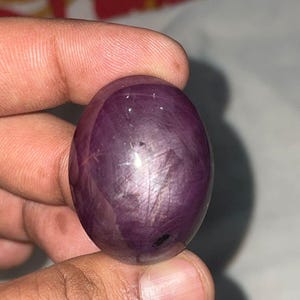 May include: A smooth, oval gemstone in a deep purple colour. The stone has a polished surface with light reflections. The gemstone is held between fingers, showing its size and colour.