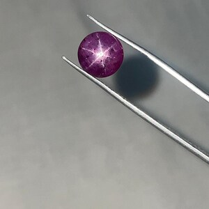 May include: A round, deep red gemstone with a six-rayed star pattern, held by silver tweezers. The gem has a polished surface and is set against a gray background. The star effect is prominent, creating a unique visual appeal.