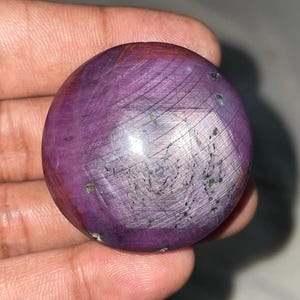 May include: A polished, round gemstone with a deep purple hue, featuring intricate surface patterns and subtle color variations. The stone is held in a hand, showcasing its smooth, spherical form and natural beauty.