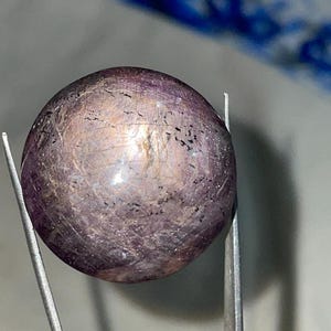 May include: A polished, spherical gemstone with a mix of purple and brown hues. The gem is held by silver tweezers, showcasing its round shape and textured surface. The stone has small dark inclusions and a subtle sheen.