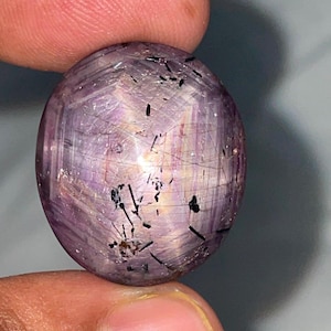 Può includere: Una gemma cabochon di forma ovale con una tonalità traslucida viola chiaro e rosa. La pietra presenta un distinto effetto a stella a sei punte ed è caratterizzata da numerose inclusioni scure a forma di ago sulla sua superficie.
