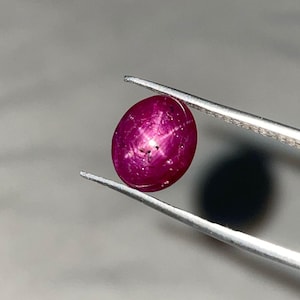 Puede incluir: Una gema de rubí ovalada, sujeta con pinzas. El rubí es de color rojo intenso y muestra un efecto de estrella de seis rayos. La gema es lisa y pulida, reflejando la luz. El fondo es de un gris suave.