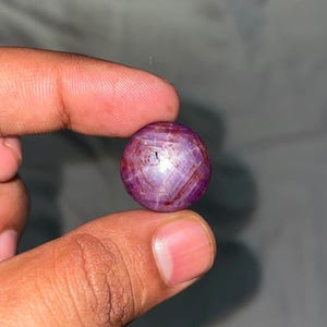 Op de afbeelding: Een ronde, in cabochon geslepen sterrobijn-edelsteen met een diepe paarsrode kleur. De steen vertoont een duidelijk zesstralig ster-effect, bekend als asterisme, zichtbaar over het gepolijste, doorschijnende oppervlak met natuurlijke insluitingen.
