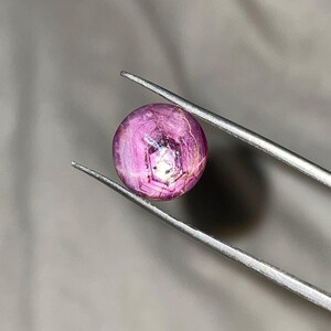 May include: A round, polished gemstone, held by silver tweezers. The gem is a deep pink color with internal striations and a bright, reflective center. The background is a neutral gray.