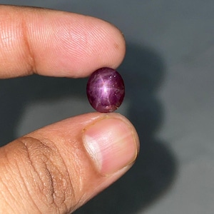 May include: A polished, oval-shaped, star ruby gemstone. The gem is a deep reddish-purple colour with a six-rayed star pattern visible on its surface. The ruby is held between two fingers.