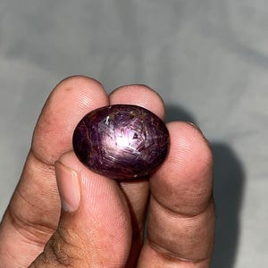 Op de afbeelding: Een ovaalvormige cabochon robijn edelsteen met een dieprode tot paarse tint. De steen vertoont een duidelijk zesstralig sterpatroon, bekend als asterisme, zichtbaar op het gepolijste, gewelfde oppervlak dat het licht subtiel weerkaatst.