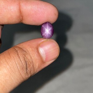 Può includere: Una piccola gemma ovale, rosso-violacea, con un motivo a stella a sei raggi. La gemma è tenuta tra due dita su uno sfondo neutro. L'effetto stella è prominente, creando un'immagine accattivante.