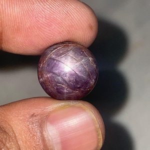 Può includere: Una gemma rotonda e lucidata, di colore viola intenso. La superficie presenta un motivo a stella e sottili linee dorate. La gemma è tenuta tra due dita, mostrando la sua forma sferica e i suoi intricati dettagli. La gemma sembra essere un rubino stellato.