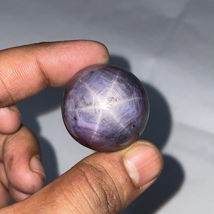 May include: A polished, spherical gemstone with a star effect. The stone is primarily a deep purple color with a six-rayed star pattern radiating from the center. The star is a lighter, almost white color. The gemstone is held between two fingers.