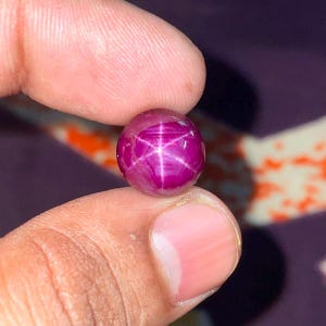 Puede incluir: Una piedra preciosa de rubí estrella de corte cabujón redondo con un color rojo magenta intenso. La piedra muestra un efecto de estrella de seis rayos, conocido como asterismo, que aparece como un patrón de luz blanca centrado.