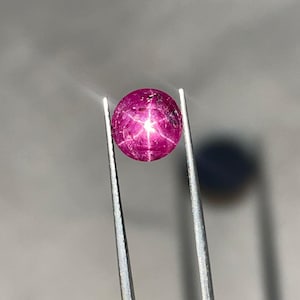 Könnte beinhalten: Ein polierter, ovaler Rubin-Edelstein, der von einer Pinzette gehalten wird. Der Edelstein hat eine tiefrosa Farbe mit einem sechsstrahligen Sterneffekt auf seiner Oberfläche. Die Pinzette ist silberfarben und der Hintergrund ist ein neutrales Grau.