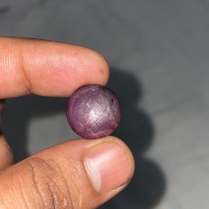 May include: A round, cabochon-cut star ruby gemstone with a deep purplish-red hue. The surface displays a visible six-rayed star effect, known as asterism, created by light reflecting off internal needle-like inclusions within the stone.