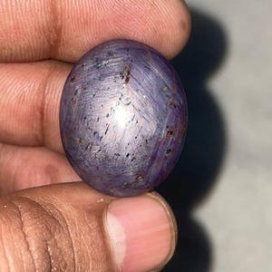 以下が含まれることがあります： 深く紫色の、磨かれた楕円形の宝石。石は星のような効果があり、中心点から線が放射状に広がっています。小さな黒いインクルージョンが宝石全体に散らばっており、そのユニークな外観を際立たせています。