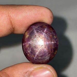 May include: A polished, oval gemstone exhibiting a star effect. The gem is a deep reddish-purple hue, displaying a six-rayed star pattern across its surface. The stone is held between two fingers against a neutral backdrop.