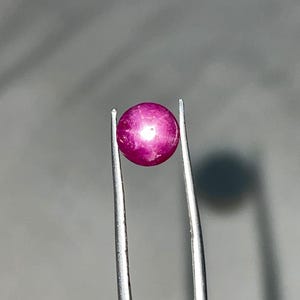 May include: A spherical, polished, deep pink gemstone held by silver tweezers. The gemstone has a smooth surface and a slight sheen, with subtle colour variations. The tweezers display the gem against a neutral background.