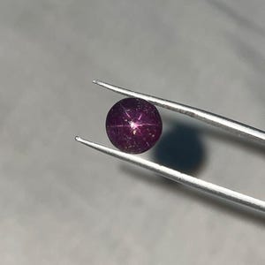Puede incluir: Una gema de rubí estrella redonda y de color rojo oscuro sujeta con pinzas. La gema muestra un patrón de estrella de seis rayos. Las pinzas son plateadas y el fondo es gris claro.