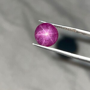 Peut inclure: Une gemme ronde, polie, rouge-violacé, avec un effet d'étoile à six rayons, tenue par une pince argentée. La gemme est d'une couleur profonde et saturée, et le motif en étoile est clairement visible. L'arrière-plan est un gris neutre flou.