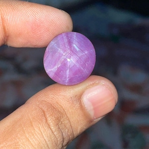 Könnte beinhalten: Ein runder, polierter Sternrubin-Edelstein. Der Edelstein hat einen leuchtenden Lilaton mit einem sechsstrahligen Sternmuster auf seiner Oberfläche. Der Stein wird zwischen zwei Fingern gehalten, wodurch seine Farbe und Form hervorgehoben werden.