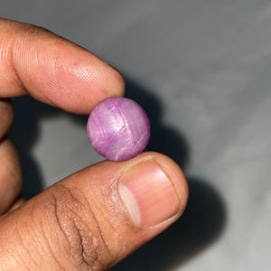 Puede incluir: Una piedra preciosa de zafiro estrella rosa, tallada en cabujón redondo, que presenta un distintivo efecto de estrella de seis puntas en su superficie. La piedra muestra un tono rosa violáceo claro translúcido con inclusiones fibrosas.