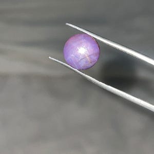 May include: A round, polished, purple gemstone with a star-like effect, held by silver tweezers. The gem has a smooth surface and a subtle color gradient. The background is a muted gray.