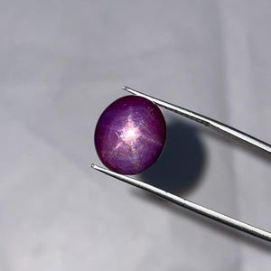 Puede incluir: Una gema pulida, de forma ovalada, de color púrpura oscuro con un efecto de estrella de seis rayos. La gema está sujeta por unas pinzas plateadas sobre un fondo gris. El efecto de estrella es visible en el centro.