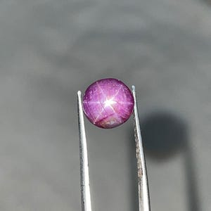 May include: A spherical, polished, purplish-red gemstone, held by silver tweezers. The gem exhibits a six-rayed star pattern across its surface. The gem is a star ruby, a type of corundum known for its asterism, the star-like effect.