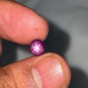 Könnte beinhalten: Ein kleiner, runder Sternrubin-Edelstein im Cabochon-Schliff mit einem tiefen Magenta-Violett-Ton. Der Stein weist einen ausgeprägten sechsstrahligen Sterneffekt auf, bekannt als Asterismus, der als heller weißer Lichtreflex auf der Oberfläche erscheint.
