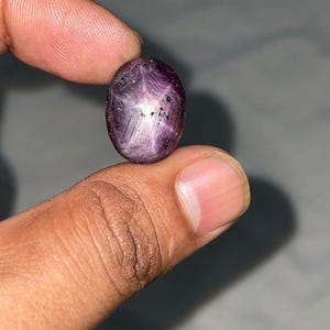 May include: An oval-shaped cabochon star ruby gemstone with a deep purple-red hue. The stone features a distinct six-rayed star effect visible on its polished surface, along with natural dark inclusions and a translucent, fibrous internal texture.