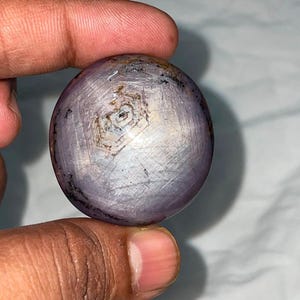 Puede incluir: Una piedra preciosa de zafiro estrella pulida y esférica que presenta un distintivo efecto de estrella de seis rayos en su superficie. La piedra muestra una paleta de colores translúcidos en violeta claro y gris con inclusiones naturales.