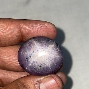 May include: A round, polished, purple gemstone with a star-like pattern. The gem is held between fingers, showcasing its spherical shape and the light reflecting off its surface. The star effect is prominent.