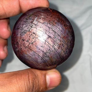 May include: A polished, spherical star ruby gemstone featuring a deep reddish-purple hue with visible natural inclusions and a distinct six-rayed star pattern across its surface, showcasing a smooth, rounded texture and reflective quality.