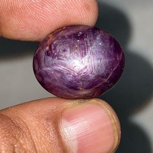 Könnte beinhalten: Ein runder, polierter Edelstein mit tiefvioletter Farbe. Der Stein weist ein sternförmiges Muster auf seiner Oberfläche auf, mit Linien, die von einem zentralen Punkt ausgehen. Der Edelstein wird zwischen zwei Fingern gehalten, um seine Größe und Form zu zeigen.