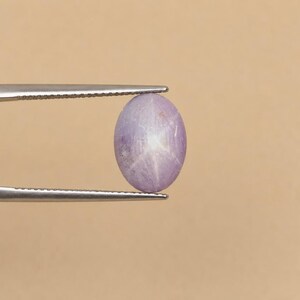 Puede incluir: Una piedra preciosa de zafiro estrella de color violeta claro, translúcida y de forma ovalada, sostenida por unas pinzas de metal plateado. La piedra presenta un efecto de estrella de seis puntas centrado en su superficie lisa y pulida.
