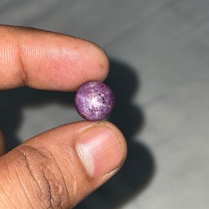 May include: A round, cabochon-cut star ruby gemstone with a deep purplish-pink hue. The stone displays a distinct six-rayed star effect, known as asterism, visible on its polished, domed surface, which features natural inclusions and dark spots.
