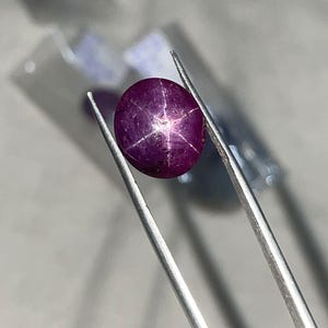 May include: A deep purple star ruby gemstone held by silver tweezers. The gemstone has a six-rayed star pattern visible on its surface. The background is blurred, with hints of other objects.