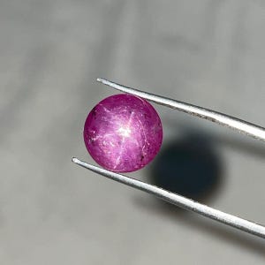 May include: A round, cabochon-cut star ruby gemstone with a vibrant magenta-pink hue. The stone displays a distinct six-rayed star effect across its polished, translucent surface, held securely by a pair of silver-toned metal tweezers.
