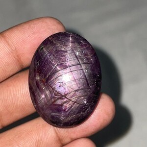 Può includere: Pietra preziosa ovale, viola intenso, con effetto stella. La superficie lucida mostra una rete di linee intersecanti, creando un motivo a stella. La gemma ha una lucentezza lucida, che riflette la luce sulla sua superficie. La pietra è tenuta in una mano.