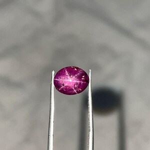 Puede incluir: Una gema de rubí pulida, de forma ovalada, sujeta con pinzas. El rubí es de color magenta intenso con un efecto de estrella de seis rayos visible en su superficie. La gema está sobre un fondo gris borroso.
