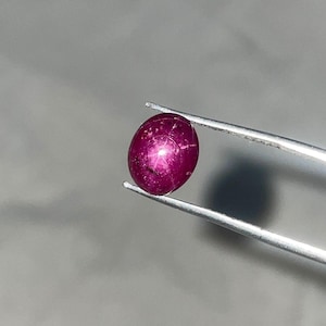 Op de afbeelding: Een diep magenta sterrobijn edelsteen met een cabochon slijpvorm, die een duidelijk zesstralig stereffect op het oppervlak vertoont. De ovaalvormige steen wordt stevig vastgehouden door een zilverkleurige metalen pincet, wat de doorschijnendheid benadrukt.