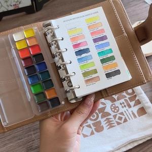 Pode incluir: Uma agenda de couro castanho contendo um conjunto de dezoito pastilhas retangulares de aguarela numa bandeja de plástico transparente e um guia de amostras de papel branco com o texto 18-COLOR WATERCOLOR SWATCH GUIDE e vários nomes de cores.