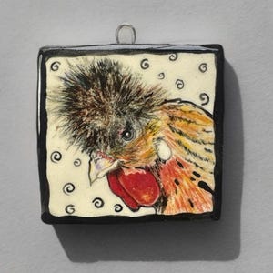 May include: A square ceramic pendant featuring a detailed illustration of a chicken with a prominent crest of dark feathers, a bright red wattle, and orange-toned plumage. The background is cream-colored with small black spiral patterns.