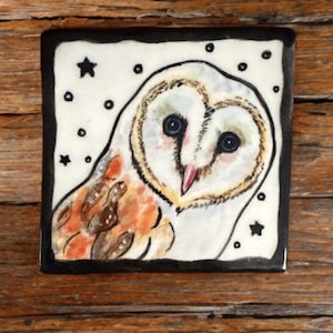 May include: Square ceramic tile featuring a hand-painted barn owl. The owl has a heart-shaped face, blue eyes, and a pink beak. The background is white with black stars and dots, framed by a black border. The tile is on a wooden surface.