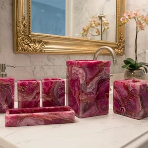 May include: A bathroom accessory set featuring a soap dispenser, two toothbrush holders, a rectangular tray, a tall wastebasket, and a square tissue box cover, all crafted from a vibrant pink and white agate stone with gold-toned accents.