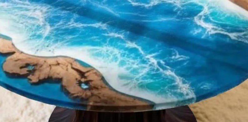 May include: Round table with a resin ocean design. The table features a vibrant blue and white wave pattern, with a section of brown, textured land. The table's design evokes a coastal theme.